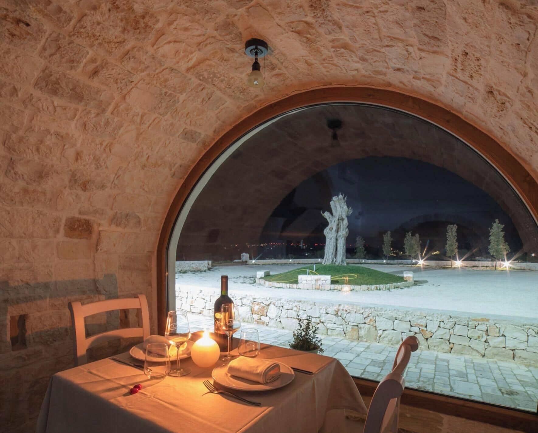 Un comedor con una mesa dispuesta para dos personas, una botella de vino y una vela. Se puede ver un parque con una escultura y árboles desde el exterior.