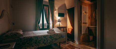 A bedroom with a bed, a bedside table, a lamp, and a bathroom visible through an open door.