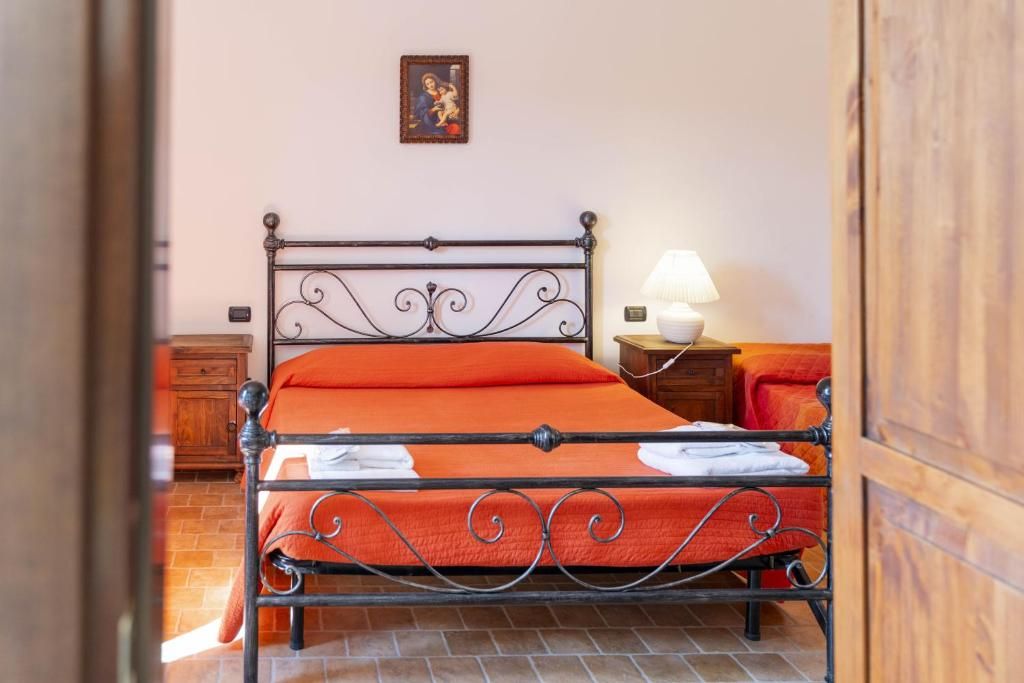 Una cama con estructura metálica y ropa de cama naranja, una lámpara de noche y una mesita de noche de madera.