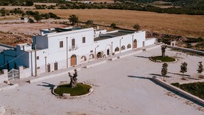 A white building with a blue section, surrounded by a paved area with small trees and a stone wall.