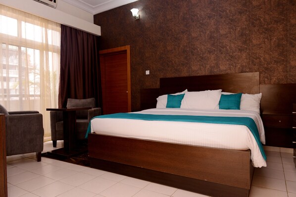 Quarto de hotel com uma cama grande, uma cadeira, uma escrivaninha e uma janela com cortinas.