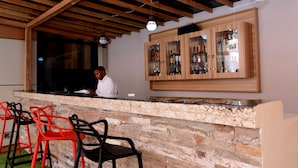 Uma área de bar com balcão de pedra, armários de madeira com garrafas e cadeiras vermelhas e pretas.