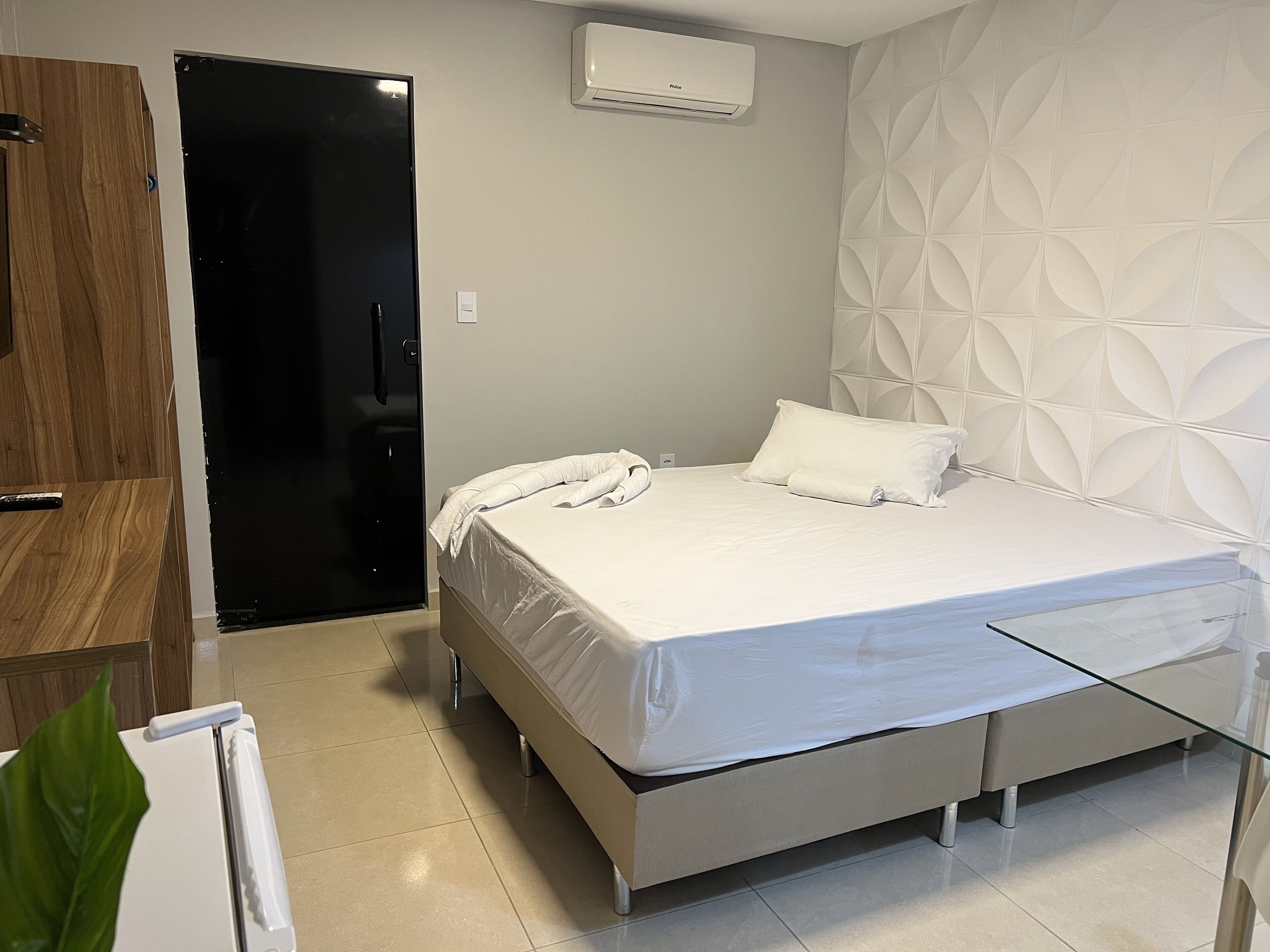 A modern hotel room with a bed, a glass table, a wooden wardrobe, and an air conditioning unit.