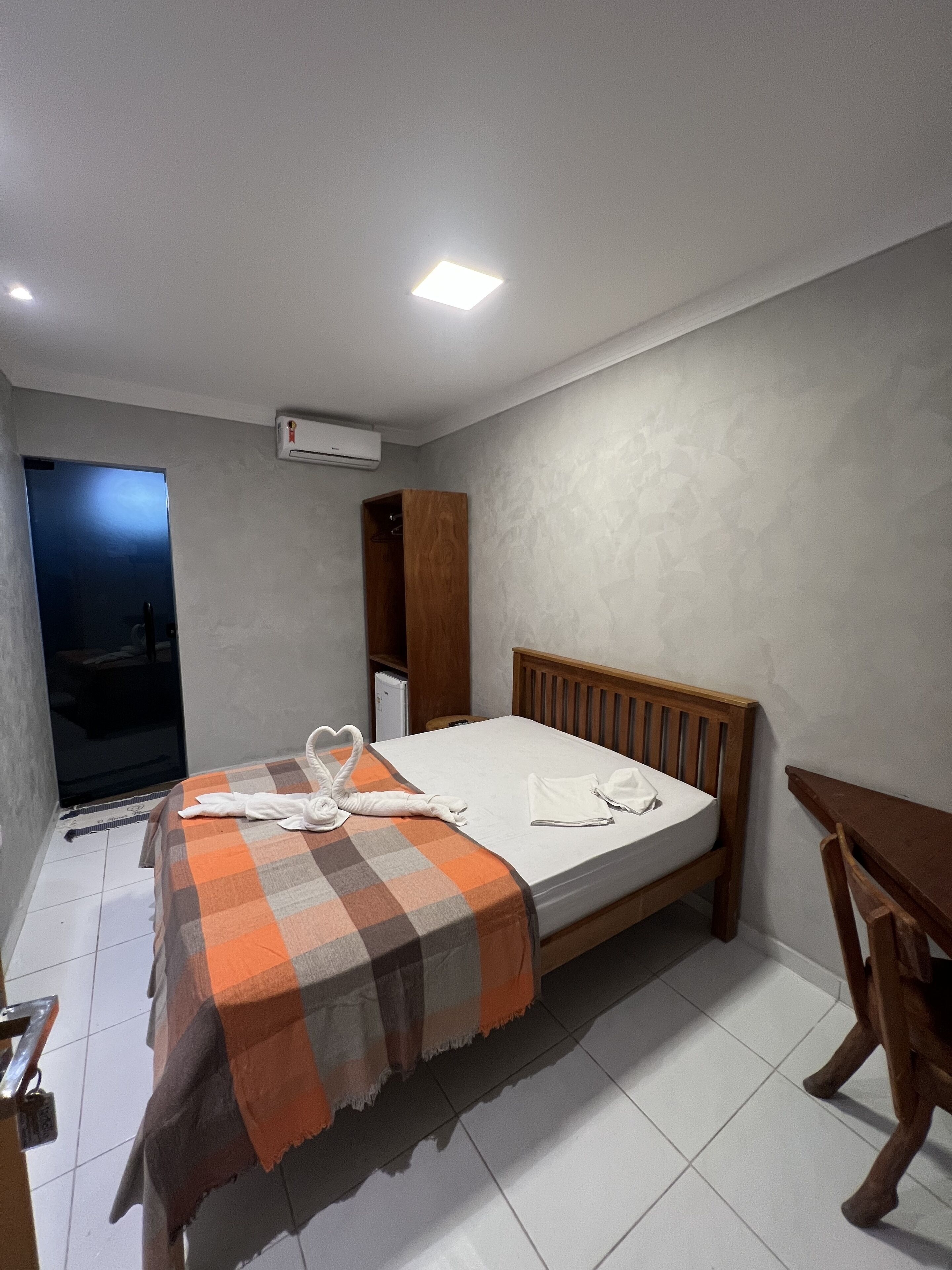 Quarto de hotel com cama, mesa de cabeceira de madeira, guarda-roupa e ar condicionado.