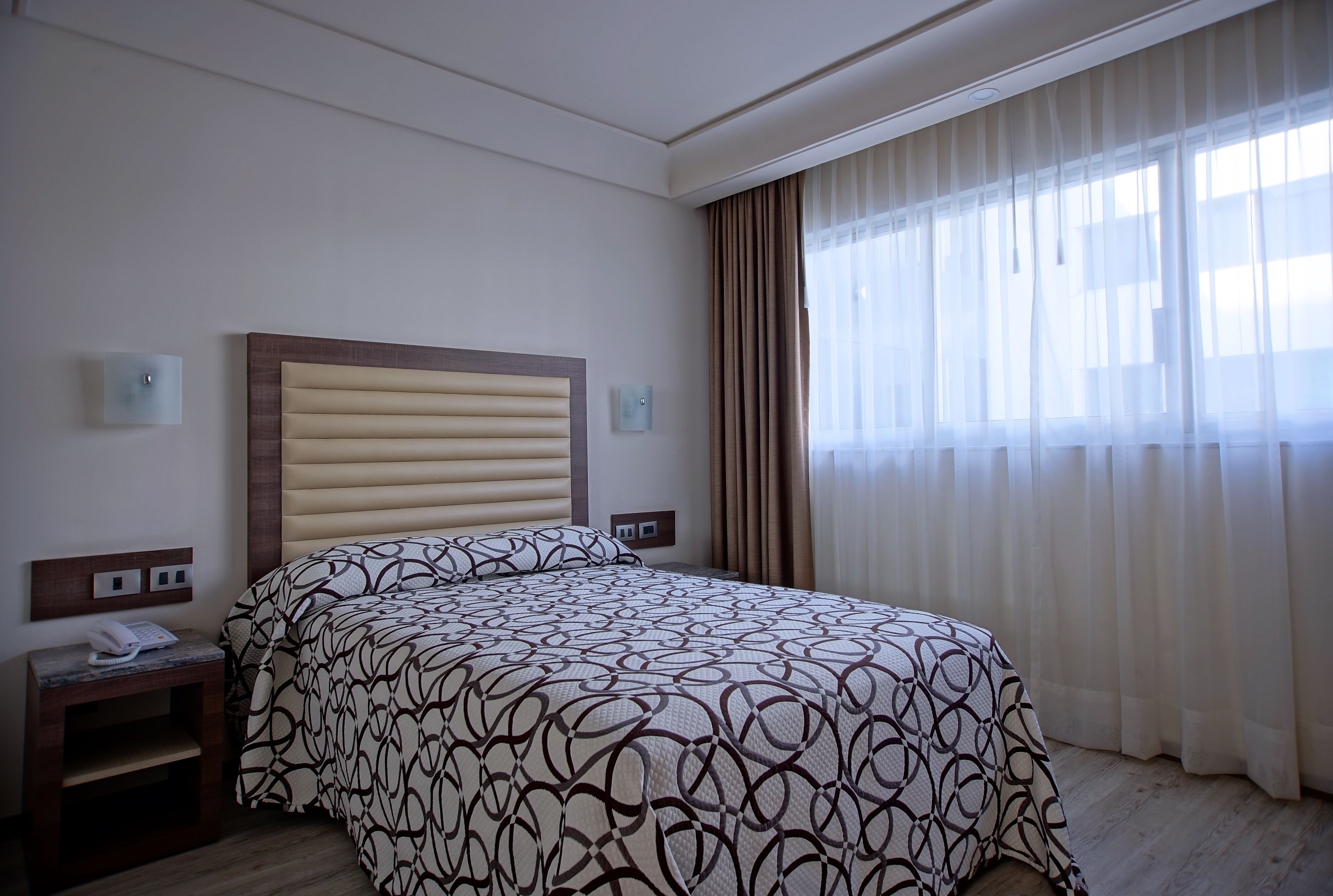 A hotel room with a large bed, a bedside table with a phone, and a window with sheer curtains.