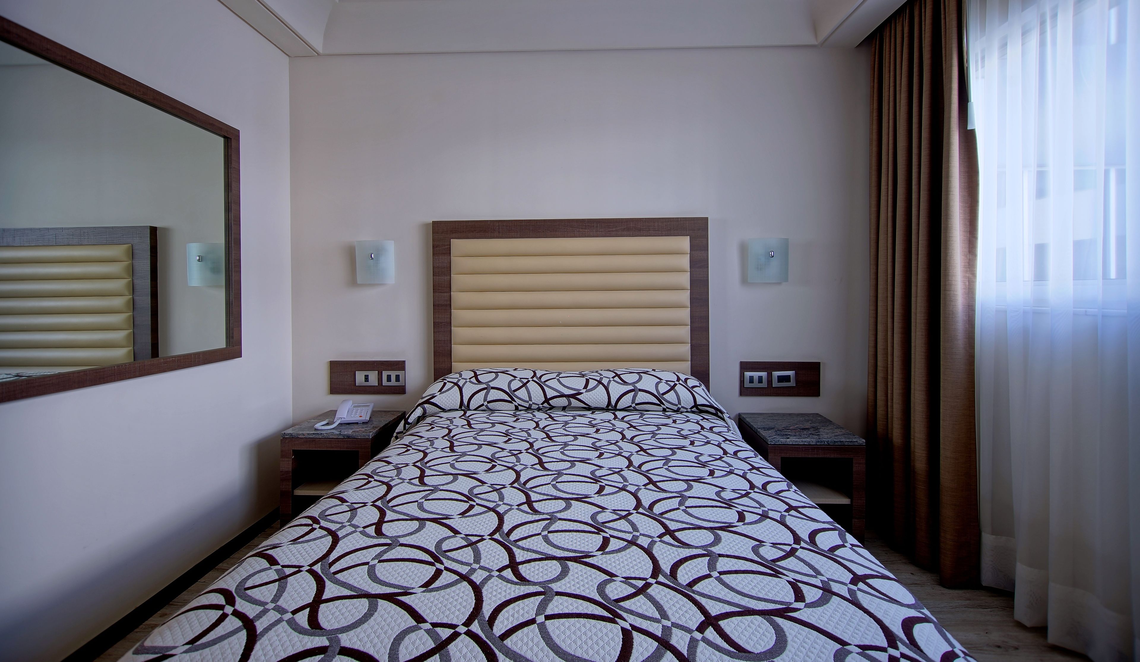 A hotel room with a large bed, a bedside table with a phone, a nightstand, a wall-mounted light, and a window with curtains.