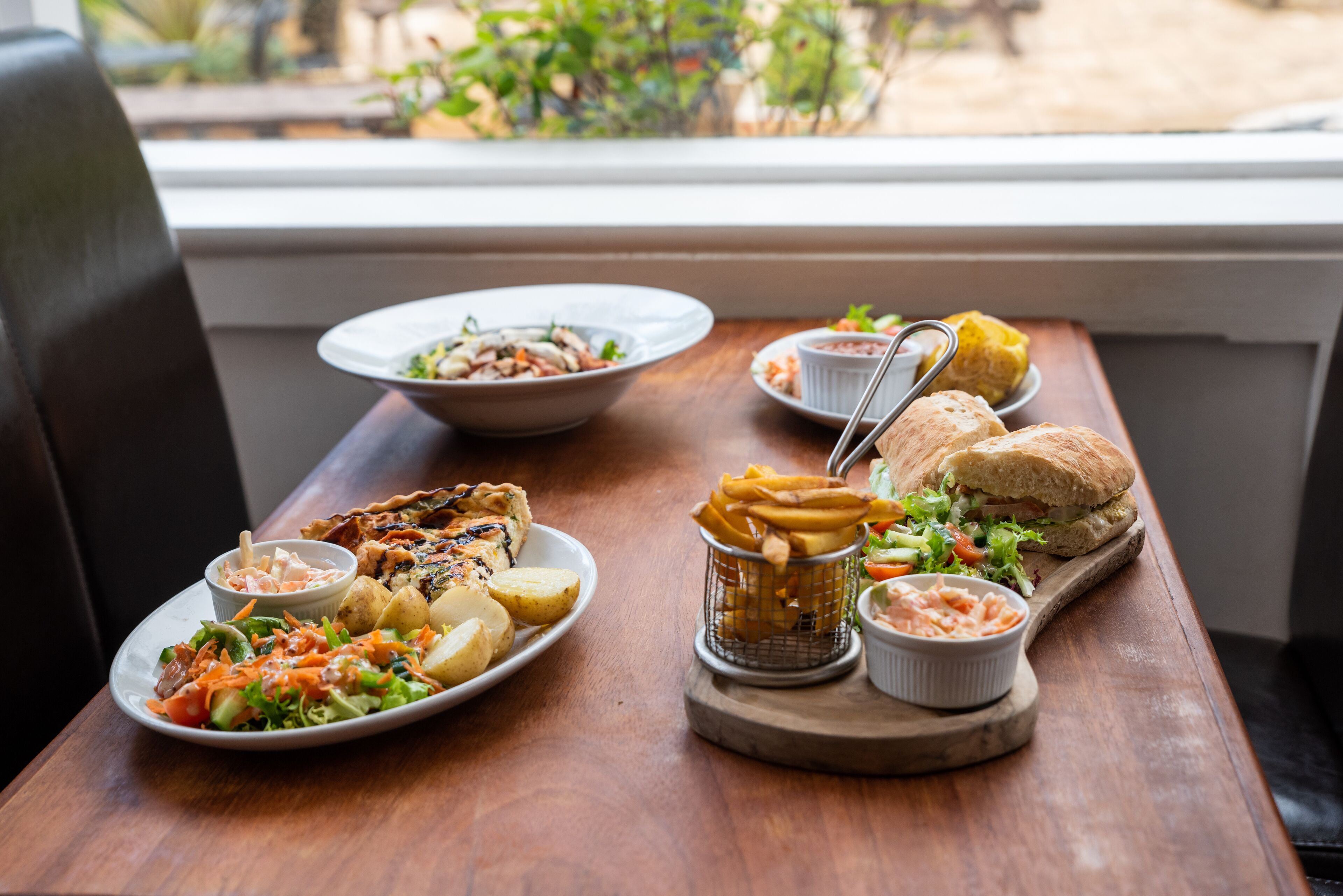 Una mesa de madera con varios platos, incluyendo una ensalada, pollo a la parrilla, papas fritas, un sándwich y un tazón de sopa.