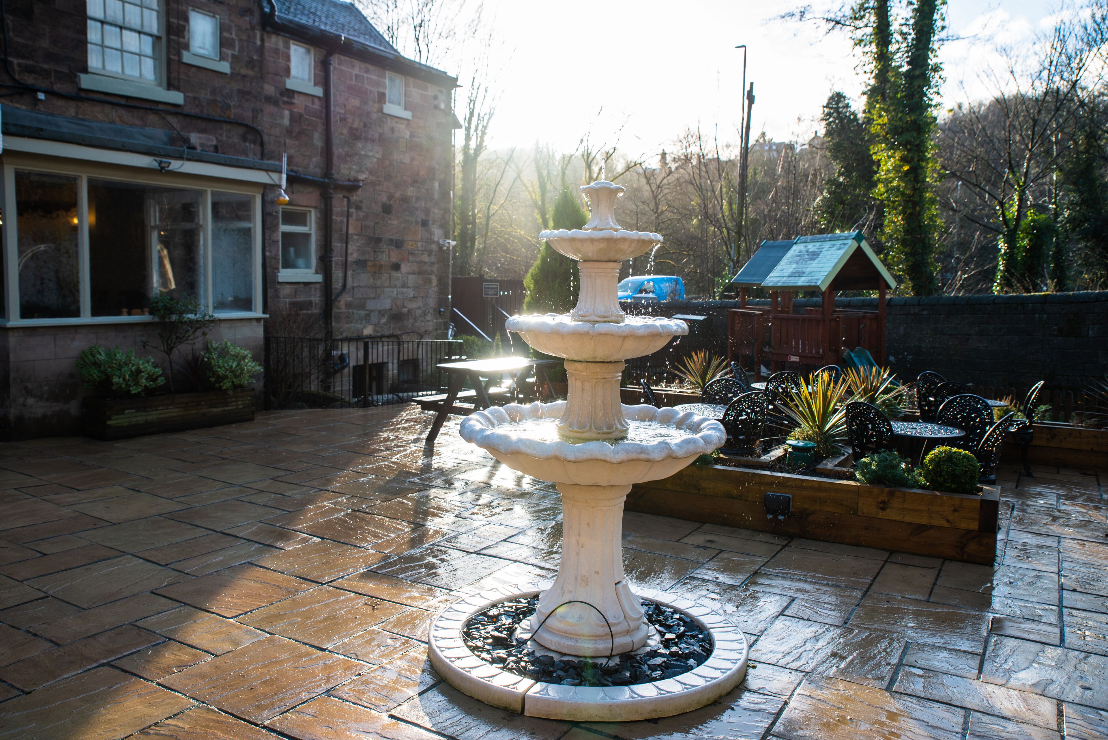 Une fontaine à plusieurs niveaux, située dans une cour pavée de pierres, et entourée de plantes en pot et de mobilier d’extérieur.