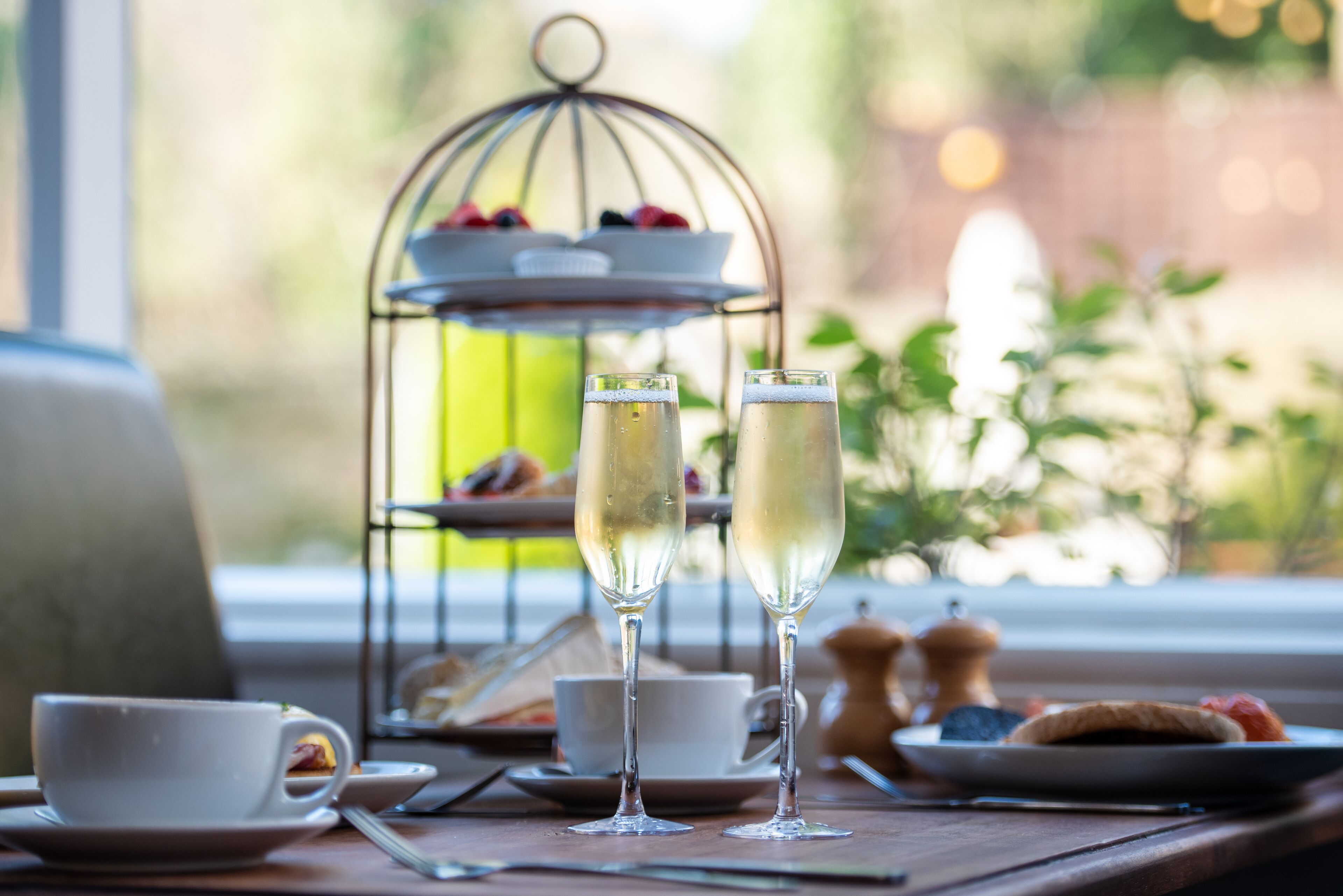 Una mesa con dos copas de champán, una taza y un soporte escalonado para postres con frutas y pasteles.