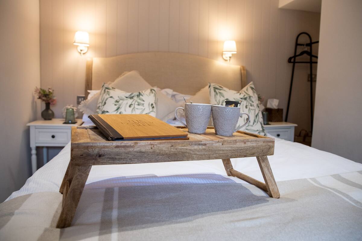 A neatly made bed with a wooden tray holding a book and two patterned cups, a bedside table with a vase of flowers, and wall-mounted lamps.