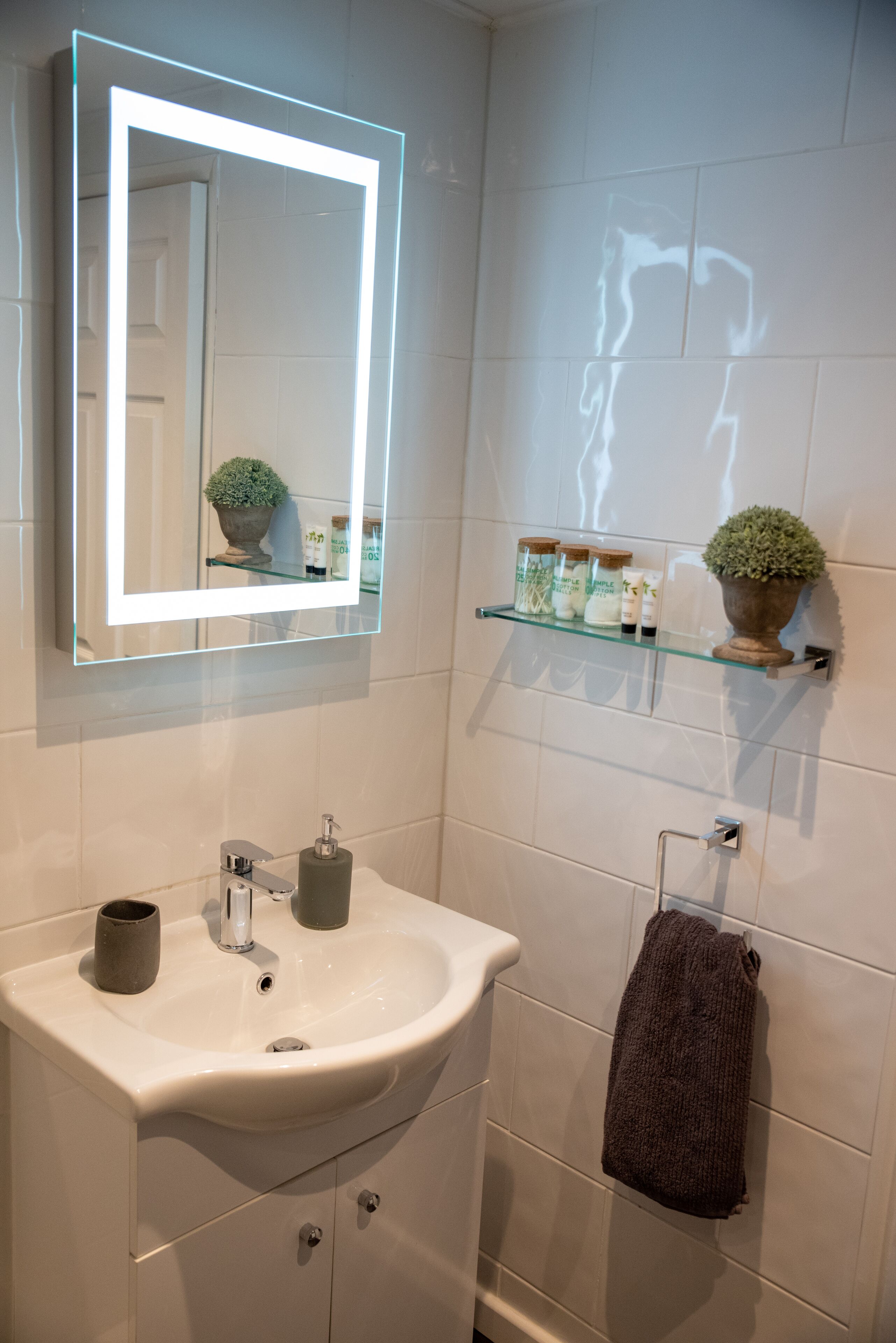 Un bagno con un lavandino bianco, uno specchio illuminato, un portascopevoli e uno scaffale con articoli da toilette.