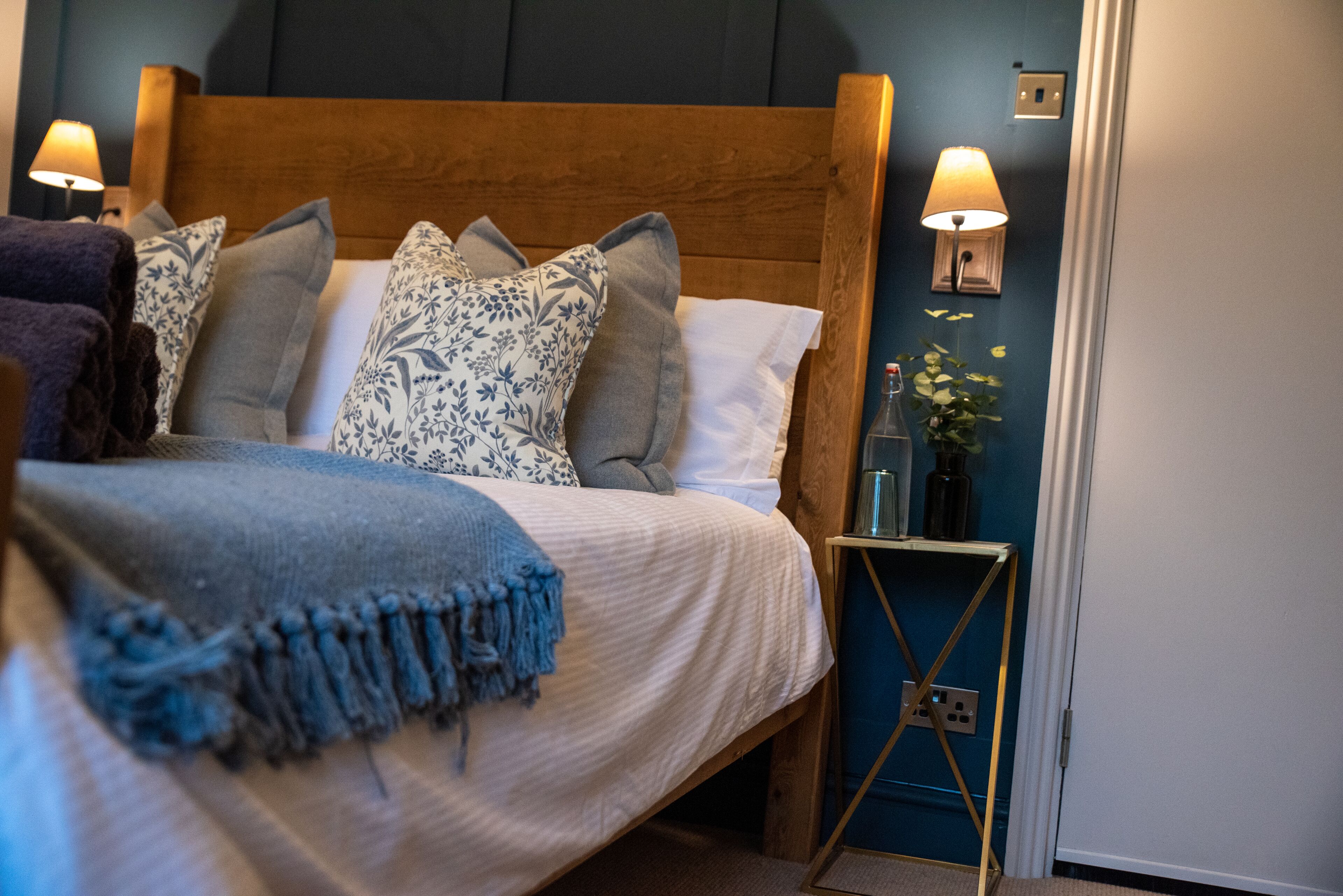 A neatly made bed with a blue throw blanket, a wooden headboard, and multiple pillows. There is a bedside table with a lamp and a small vase with flowers.