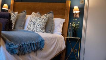 Una cama impecable con una manta azul, un cabecero de madera y varias almohadas. Se observa una mesita de noche con una lámpara y un pequeño jarrón con flores.