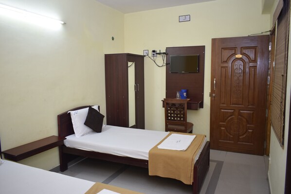A single bed with a wooden headboard, a small wooden cabinet, a television mounted on the wall, and a wooden door with a number plate.