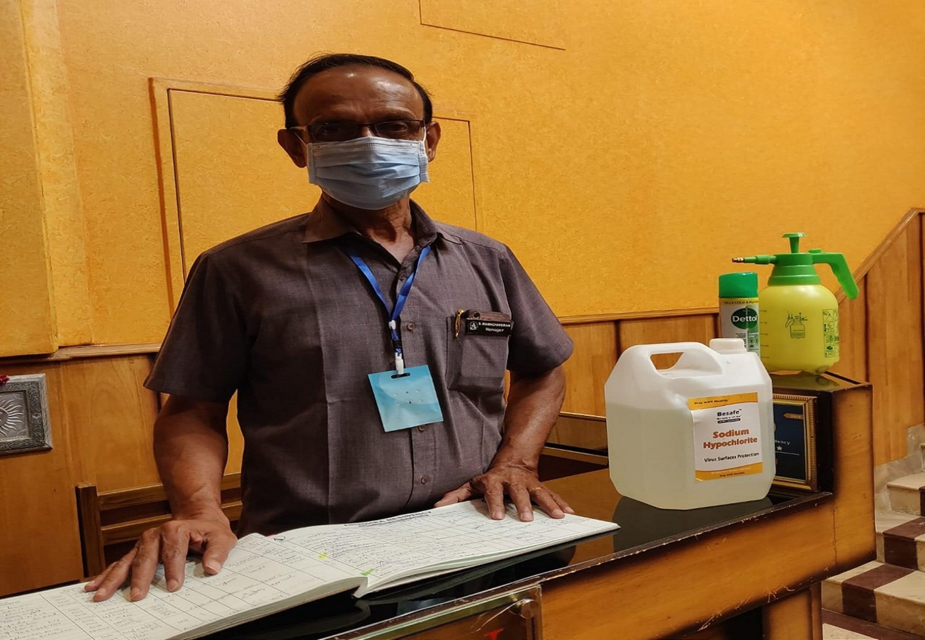 Um homem está em frente ao balcão de recepção, segurando um bloco de notas, usando crachá e máscara. Há produtos de limpeza sobre a mesa.