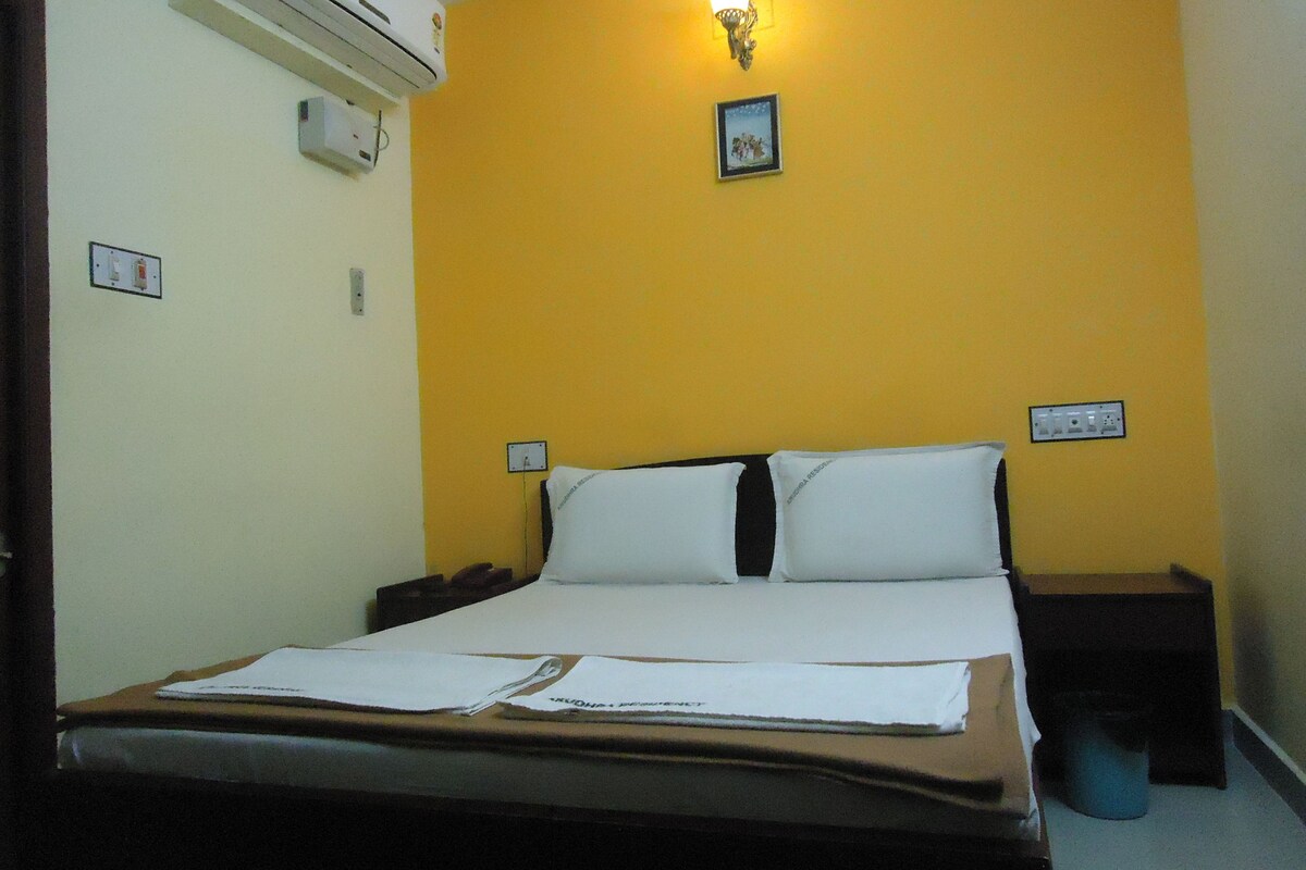 A hotel room with a bed, a nightstand, a wall-mounted air conditioner, a wall socket, and a wall-mounted light fixture.