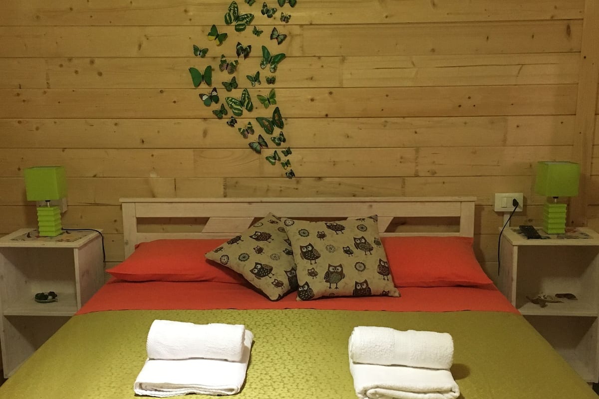 A room with a wooden wall, a bed with red and green bedding, two white towels on the bed, and a wall decoration of butterflies.