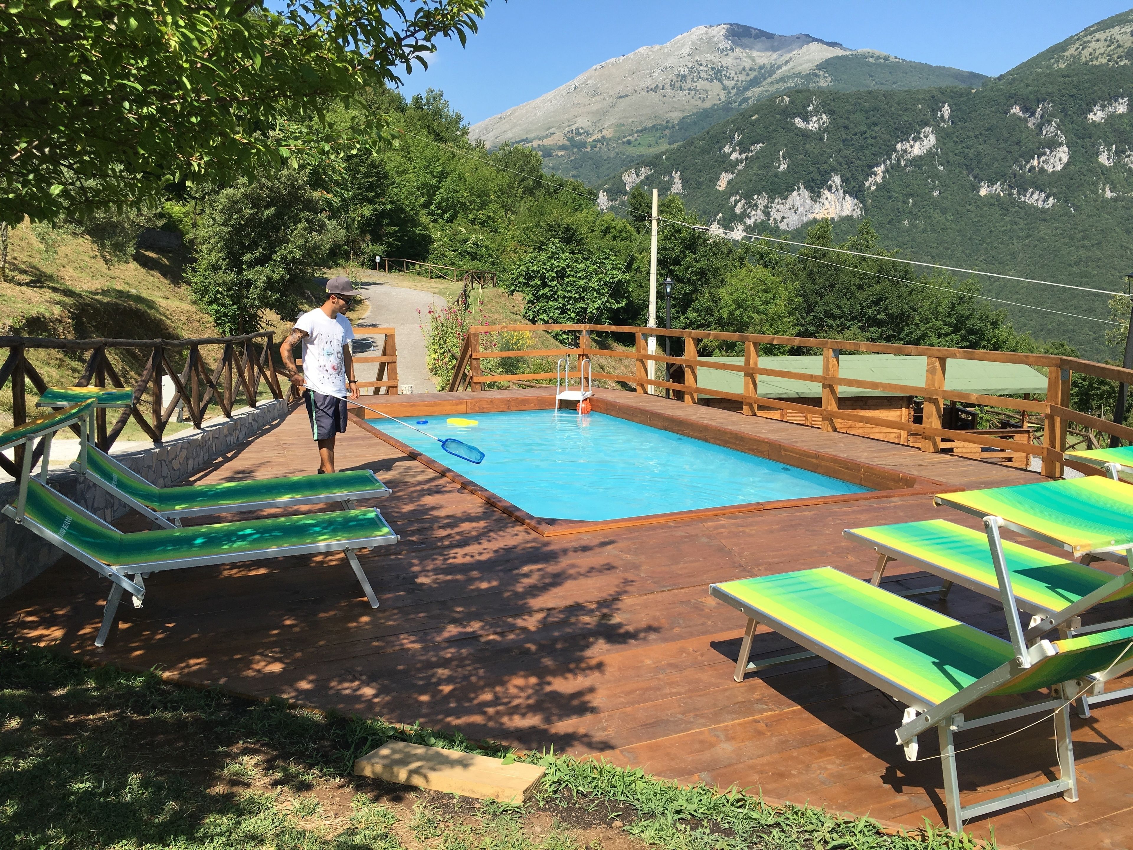 A pool area with lounge chairs and a wooden deck, surrounded by greenery and mountains.