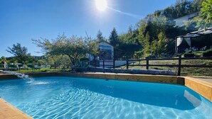 A clear swimming pool with wooden deck edges, surrounded by a fenced area with chairs and a building in the background. - B&B Lao (Papasidero)