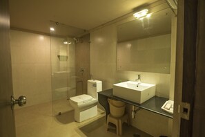 A modern bathroom with a glass shower enclosure, a white toilet, a sink with a mirror above it, and a small stool. - Hotel Nisarga (Bhopal)