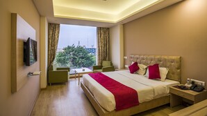 A hotel room with a bed, a desk, a chair, a TV, and a window with curtains. - Hotel Nisarga (Bhopal)
