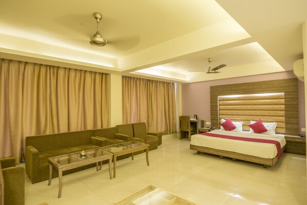 A hotel room with a sofa, a glass-top coffee table, a bed with a wooden headboard, and a ceiling fan. - Hotel Nisarga (Bhopal)
