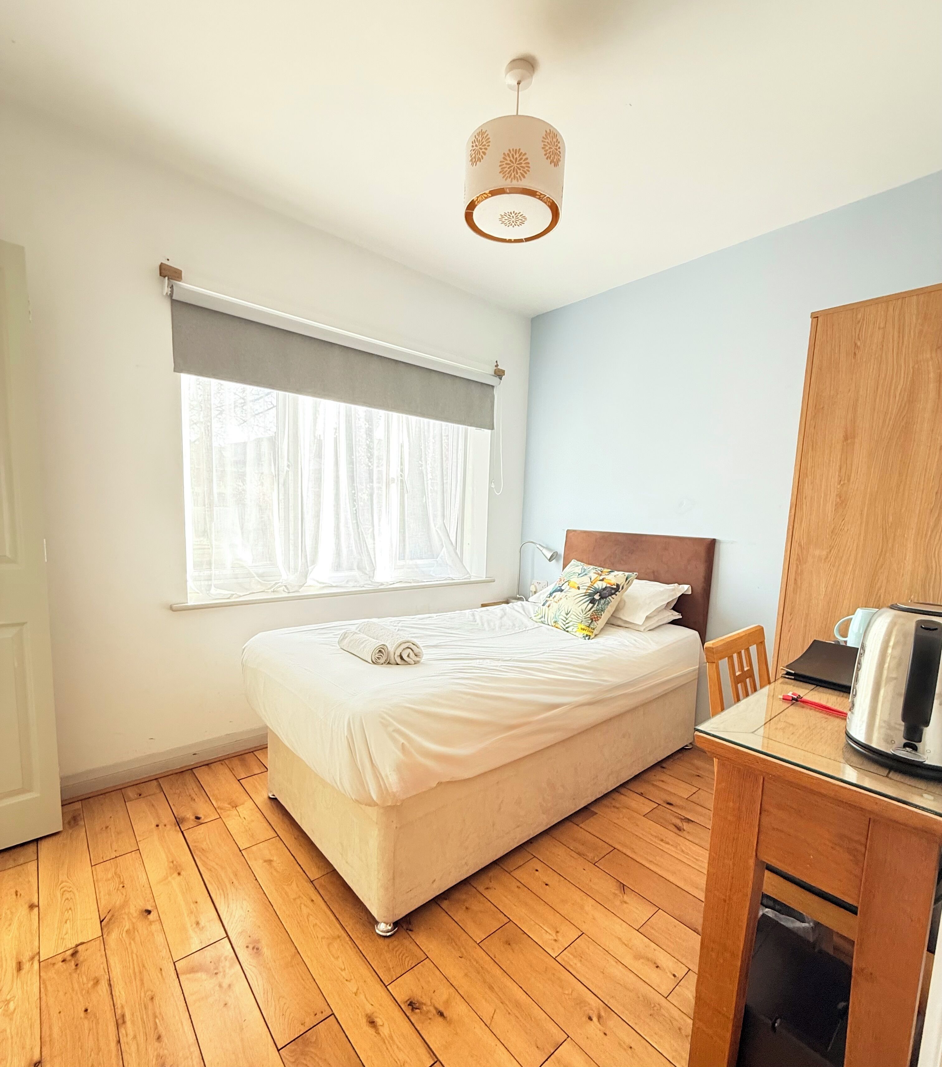 A neatly arranged bedroom with a bed, a wooden chair, a table with a kettle, and a window with curtains.