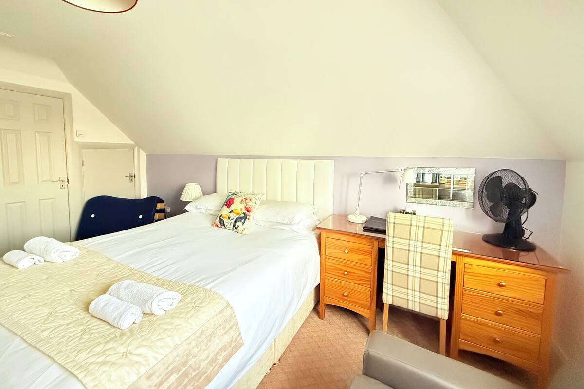 A hotel room with a bed, bedside tables, a desk, a chair, a lamp, a fan, and a window with a view.
