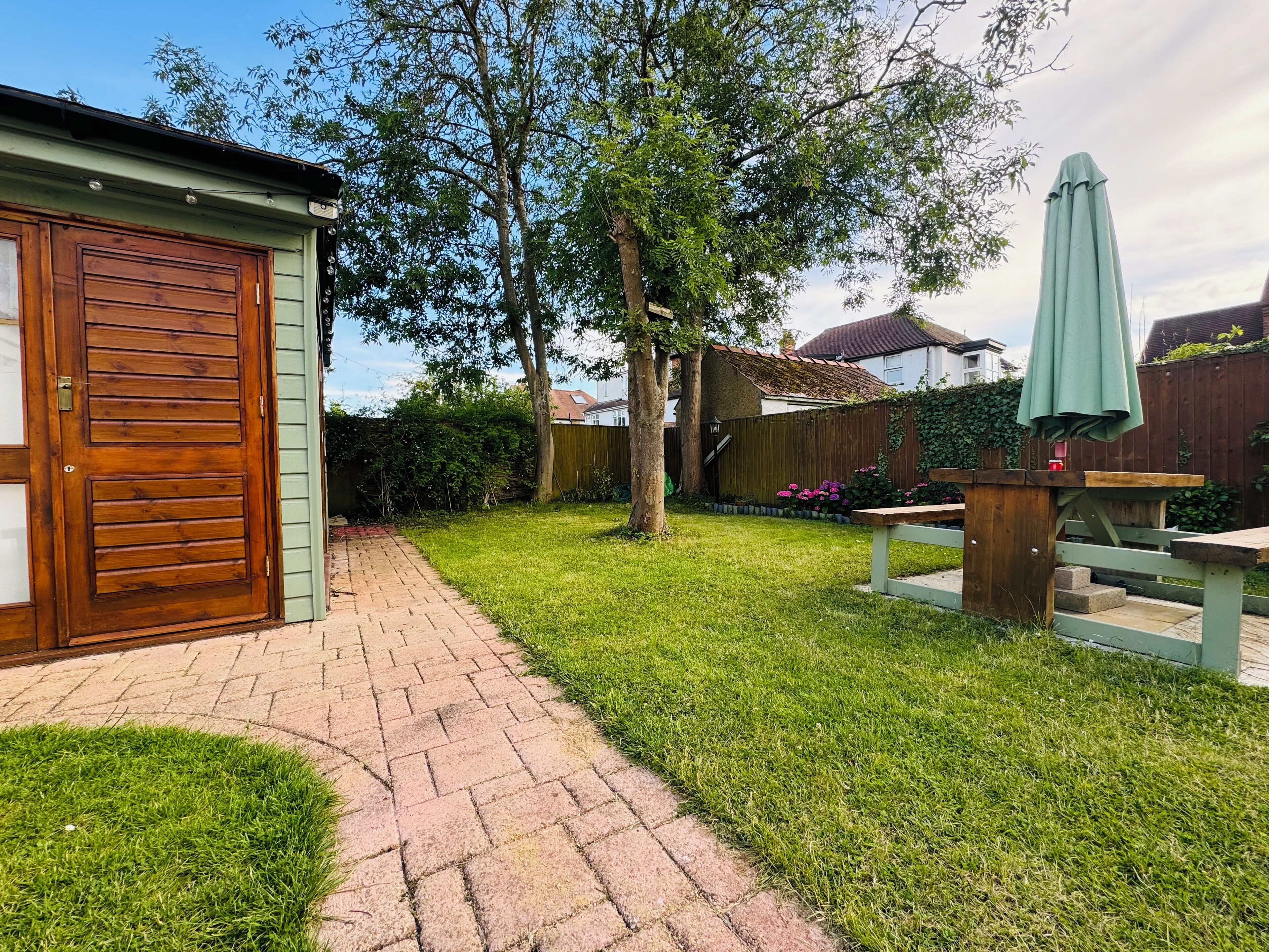 A backyard with a wooden shed, a paved walkway, a picnic table, and a green umbrella.