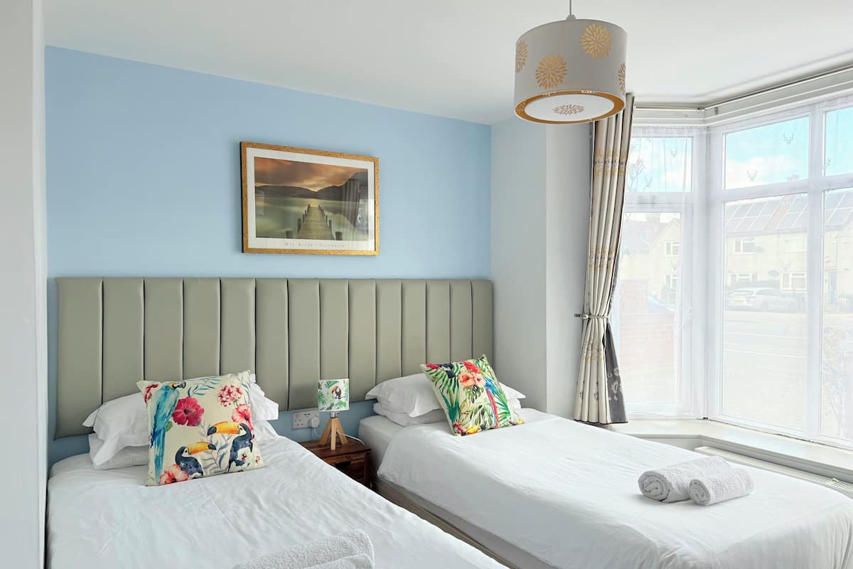 A hotel room with two beds, a large window with curtains, a ceiling light, and a framed picture on the wall.