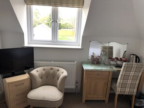 A room with a television on a cabinet, a tufted armchair, a wooden desk with a mirror, and a window with blinds.