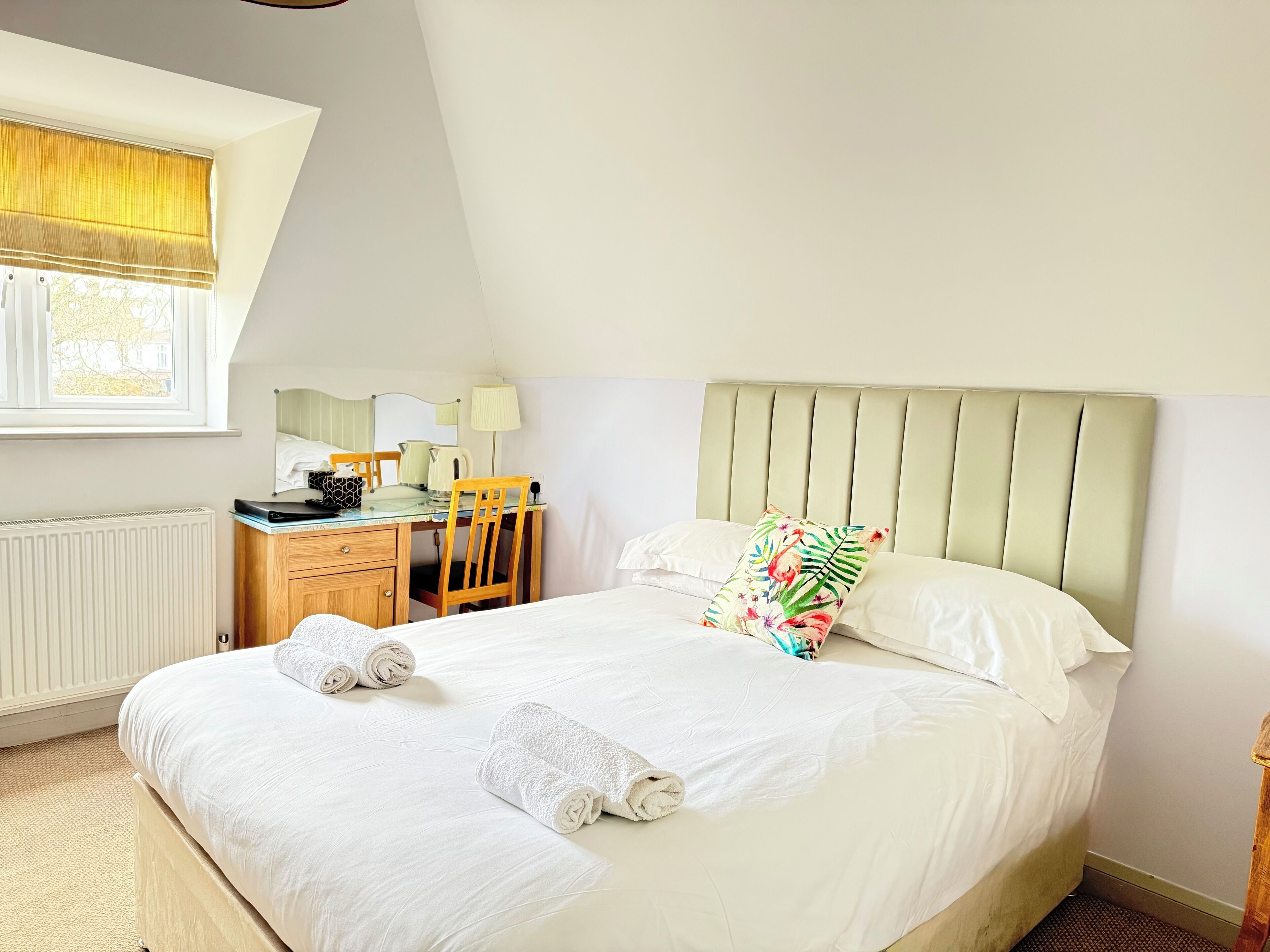A neatly made bed with white linens and two rolled towels, a wooden desk with a lamp, and a window with a yellow blind.