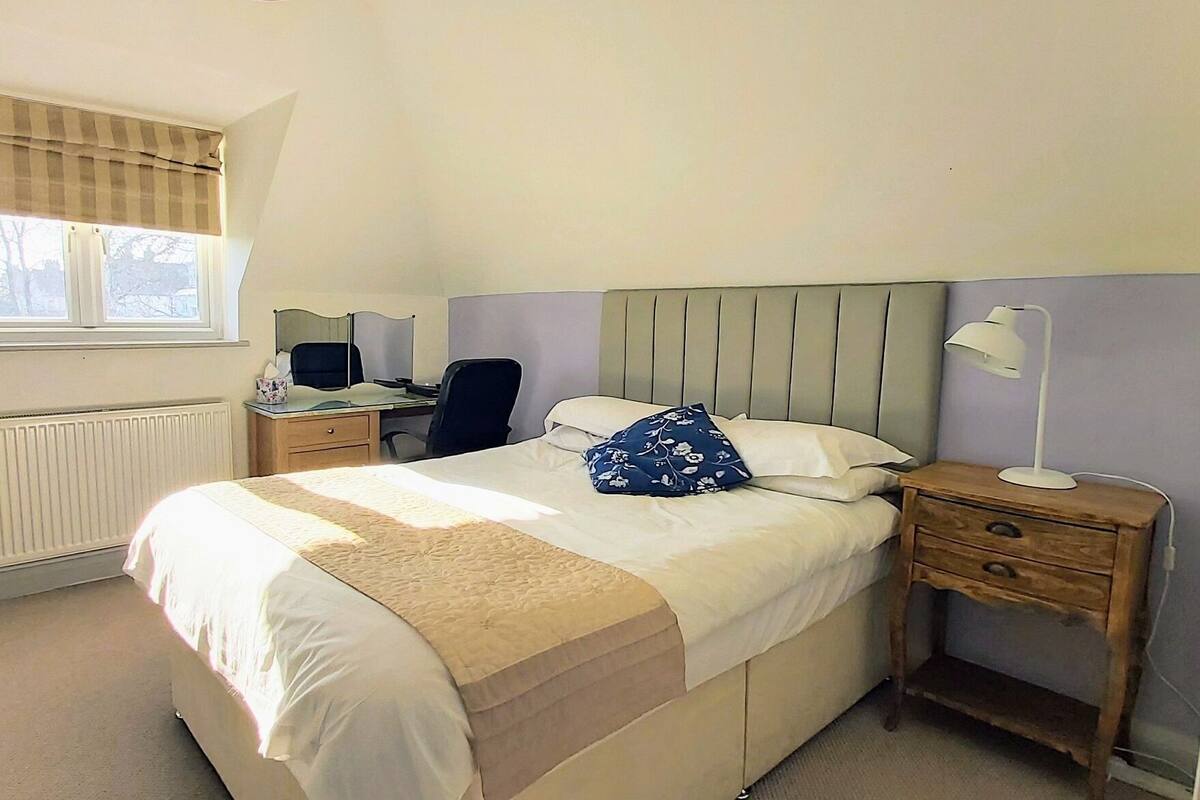 A bedroom with a bed, bedside tables, a desk, a chair, a radiator, a window with blinds, and a television on the wall.