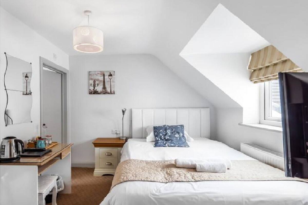 A bedroom with a sloped ceiling, a bed with a white headboard, a bedside table with a lamp, a window with curtains, and a painting on the wall.
