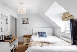 A bedroom with a sloped ceiling, a bed with a white headboard, a bedside table with a lamp, a window with curtains, and a painting on the wall.