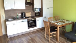 A compact kitchen with white cabinets, a built-in oven, and a dining area with a wooden table and chairs. - Ferienwohnung Mayer (Rangsdorf)