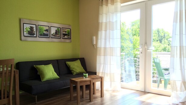 A living room with a black sofa, green cushions, a small wooden table, and a glass of green drink. There are three framed pictures on the wall and a sliding glass door leading to an outdoor area. - Ferienwohnung Mayer (Rangsdorf)