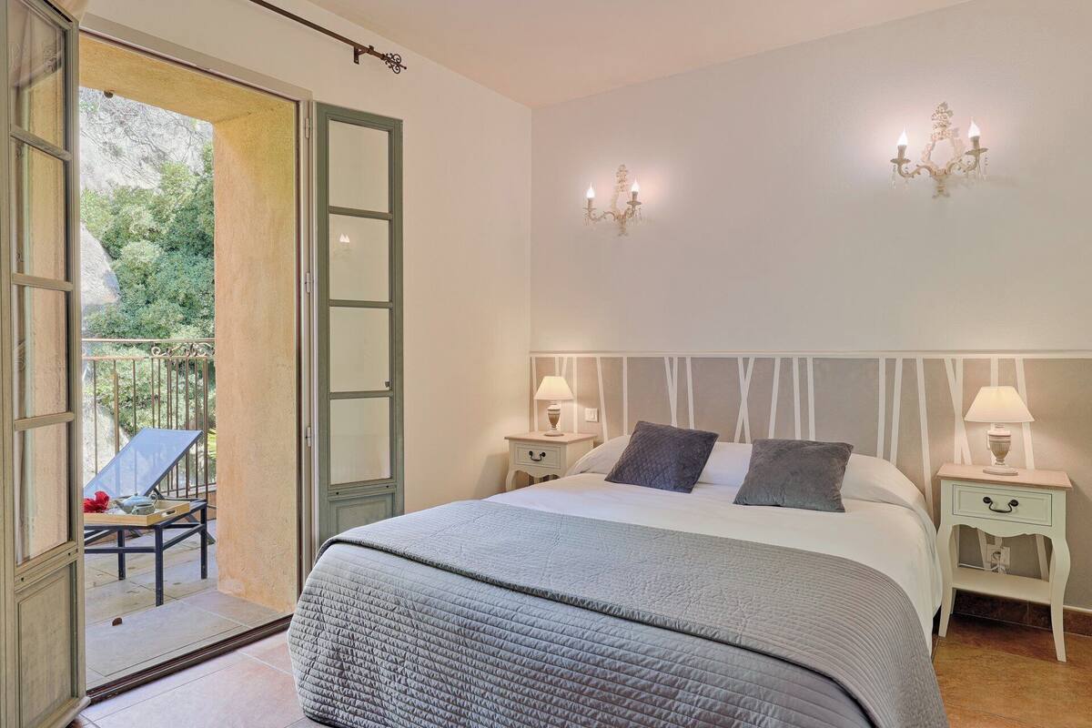 Un dormitorio con una cama grande, dos mesitas de noche con lámparas, un balcón con mesa y sillas, y vistas al verde exterior.