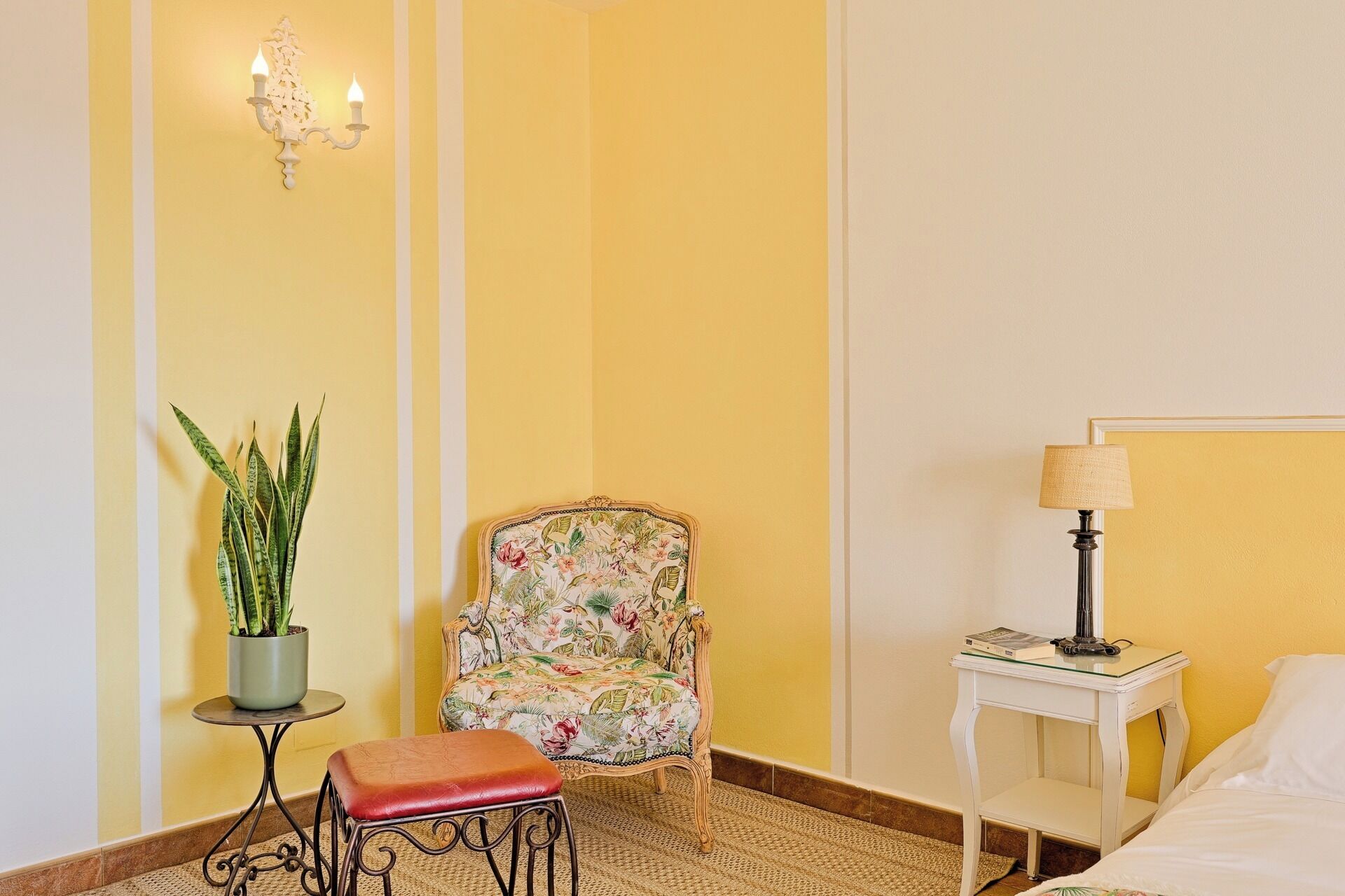 Une chambre d’hôtel avec un fauteuil à motifs floraux, une petite table sur laquelle est posée une plante en pot, une table de chevet avec une lampe et un lit.