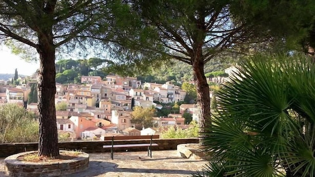 A stone-paved area with a bench, surrounded by trees and a view of a densely packed town with red-roofed buildings. - Hostellerie du Cigalou (Bormes-Les-Mimosas)