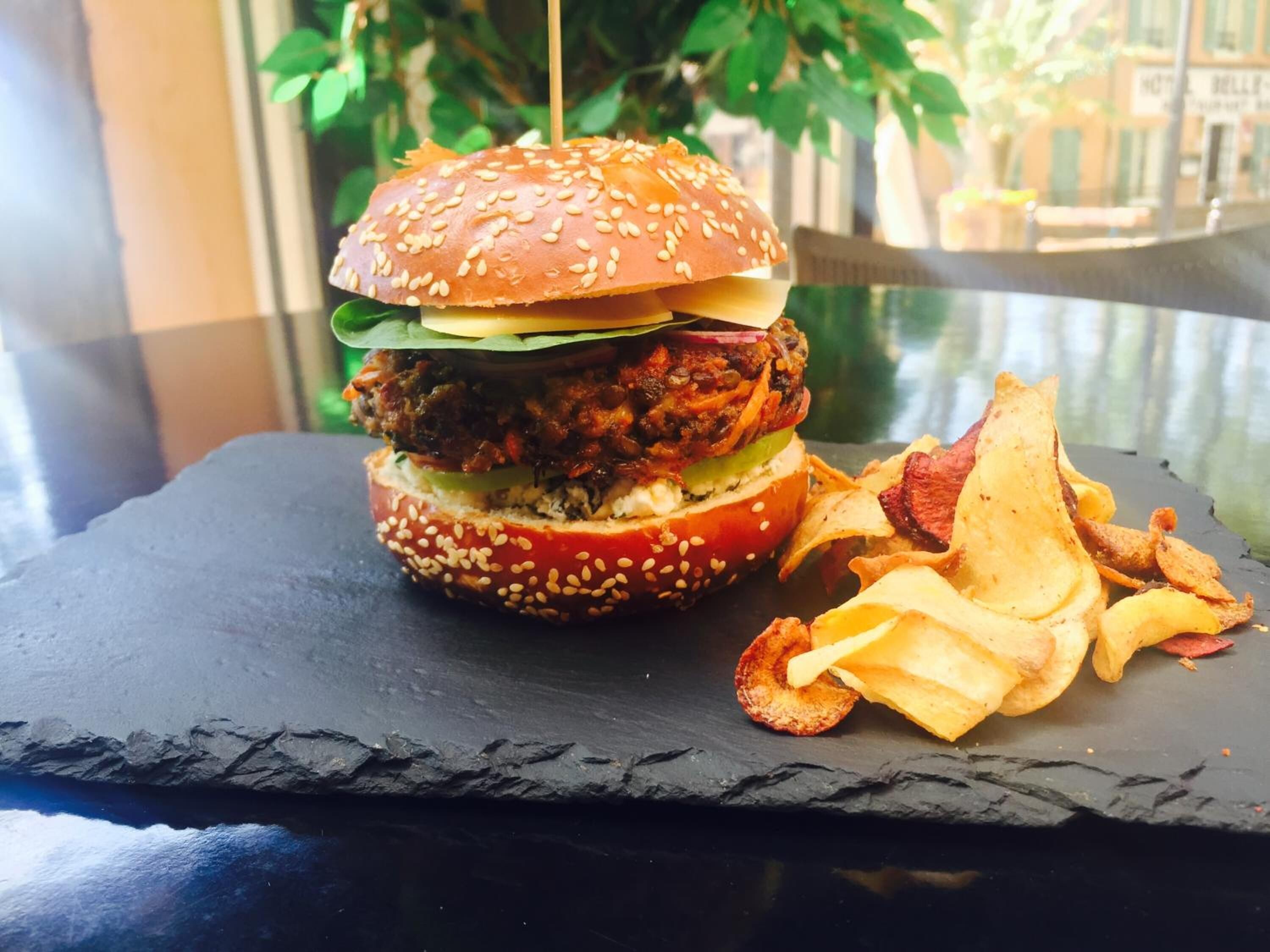 A burger with a sesame seed bun, lettuce, and a meat patty, served with potato chips on a slate plate.
