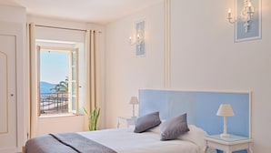 A bedroom with a large bed, two bedside tables with lamps, a view of the sea through a window, and a balcony with a railing. - Hostellerie du Cigalou (Bormes-Les-Mimosas)