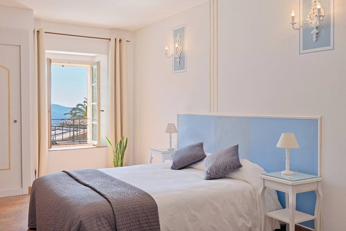 Un dormitorio con una cama grande, dos mesitas de noche con lámparas, vista al mar a través de una ventana y un balcón con barandilla.
