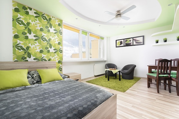 A bedroom with a bed, a desk, two chairs, a ceiling fan, and a window with blinds.