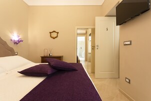 Una habitación de hotel con cama, mesita de noche con lámpara, televisor montado en la pared y una puerta que da a otra habitación.