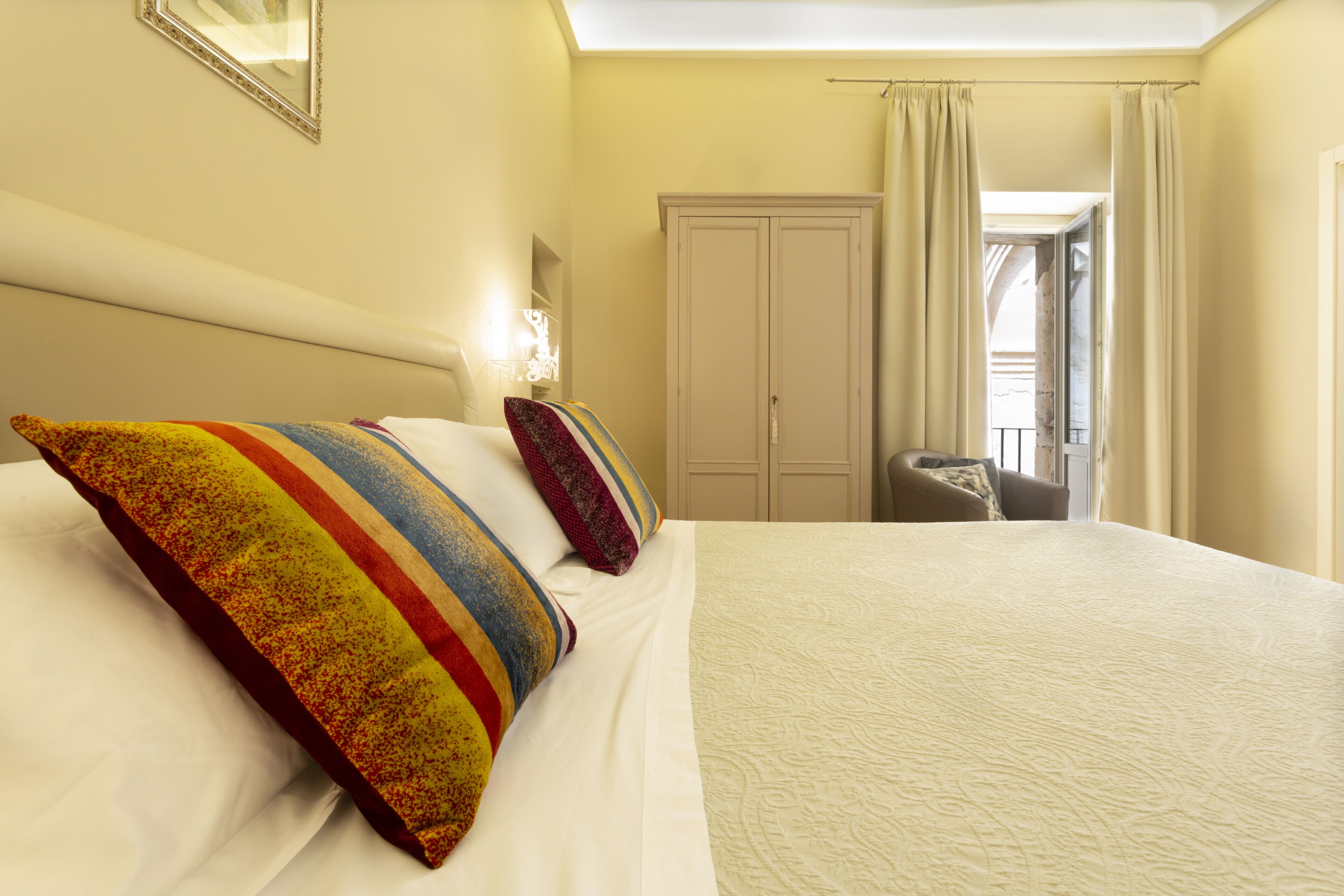 A hotel room with a large bed, two pillows, a nightstand, a lamp, a wardrobe, and a view of the outside.