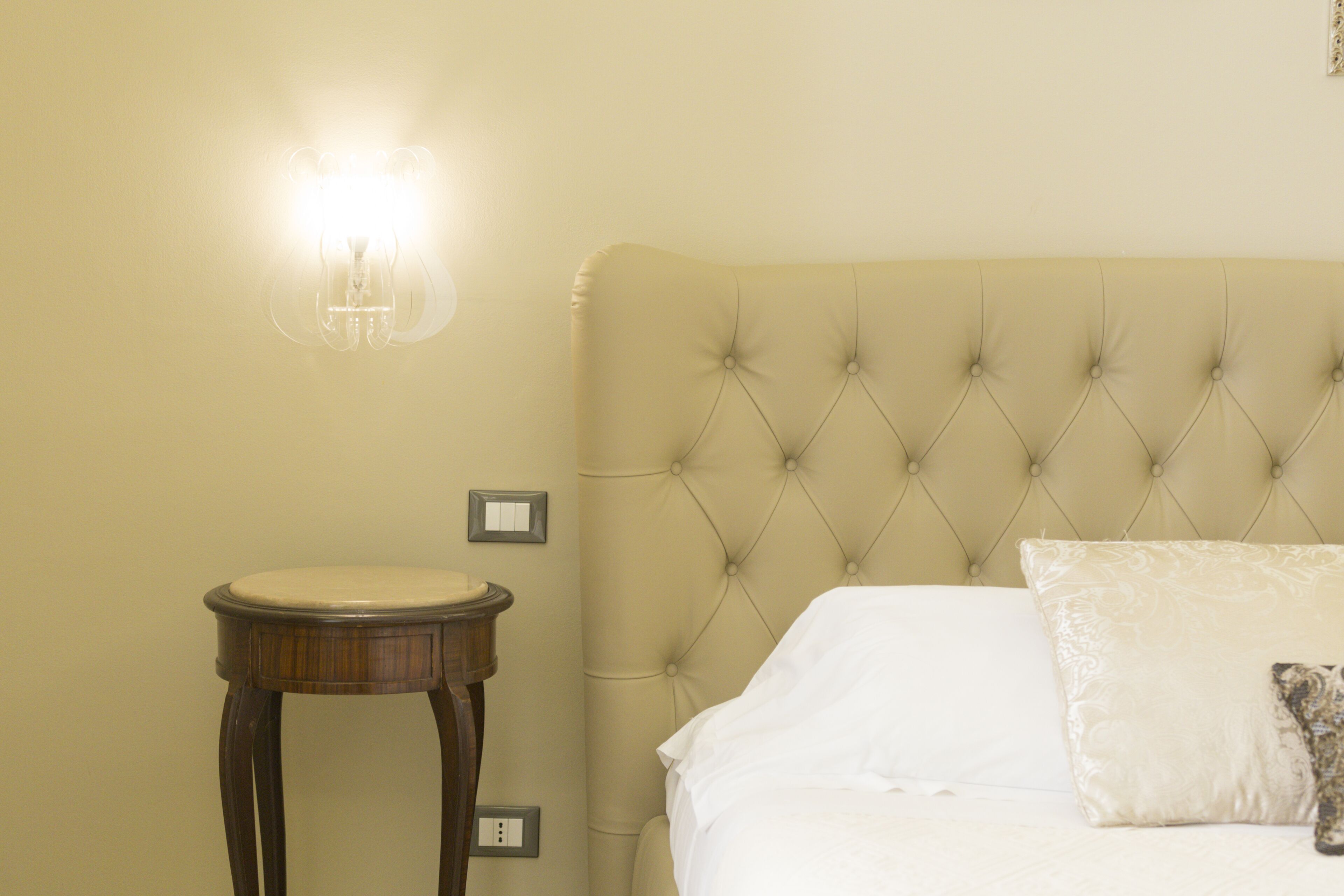A bedroom with a tufted headboard, a round wooden nightstand, and a wall-mounted light fixture.