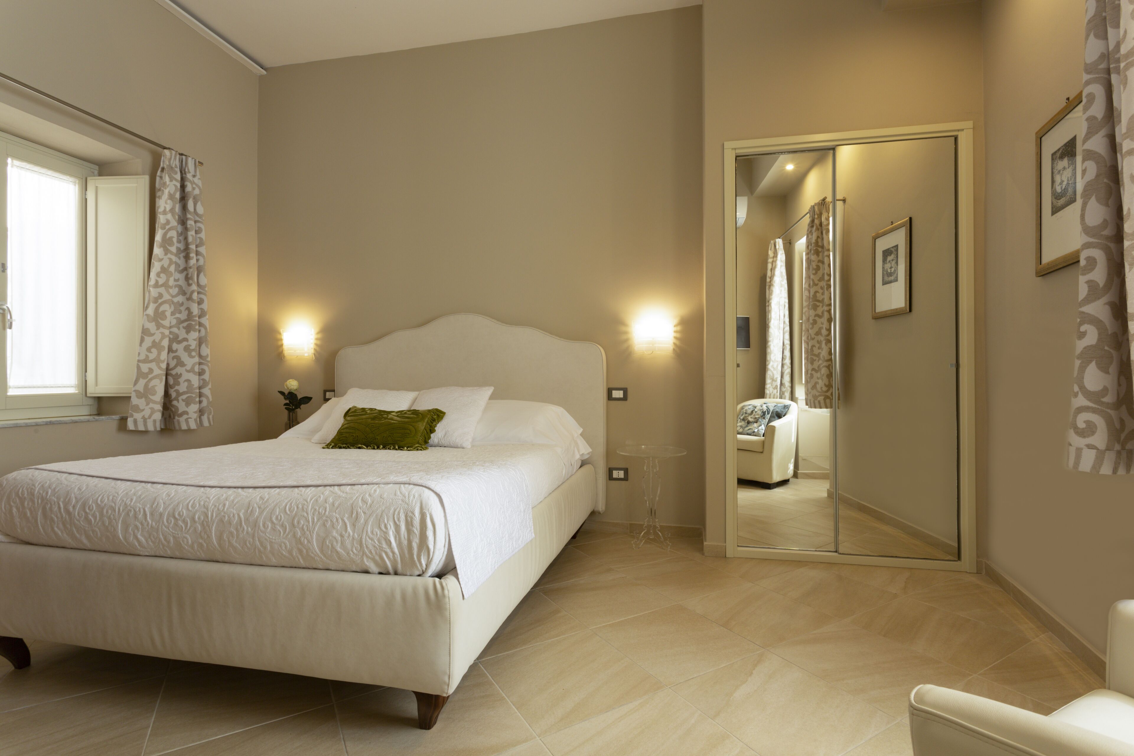 A bedroom with a bed, bedside lamps, a mirror, and a door leading to another room.