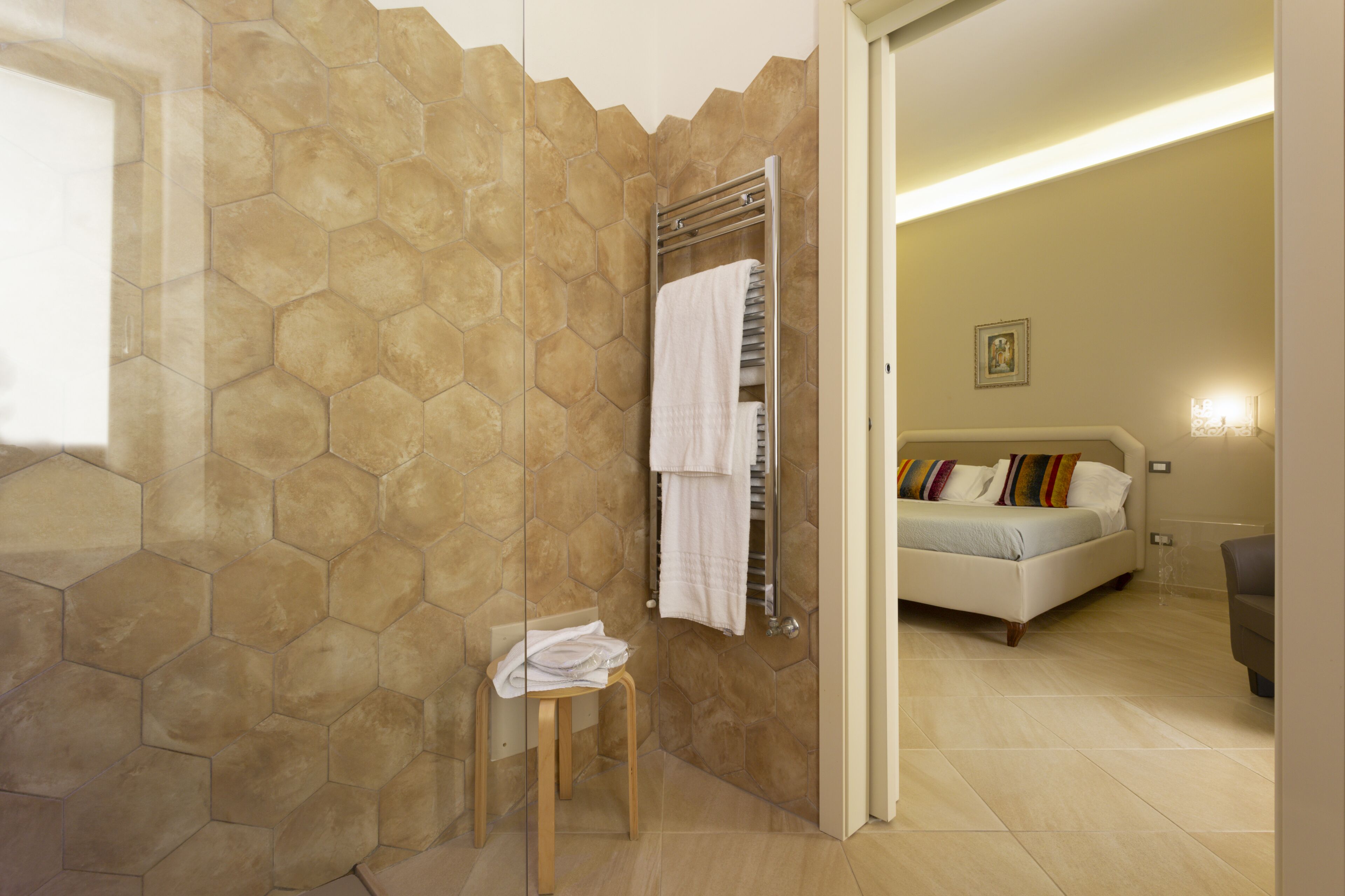 Ein Badezimmer mit Wand aus sechseckigen Fliesen, einem Handtuchhalter mit Handtüchern, einem hölzernen Hocker und einem angrenzenden Raum mit einem Bett und Kissen.