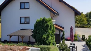 Un bâtiment à deux étages, doté d’un toit en pente, d’un patio couvert et d’un petit jardin avec un banc et un parasol.